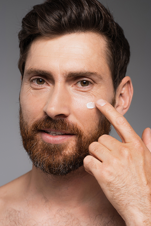 close up of man with beard applying cream on face isolated on grey