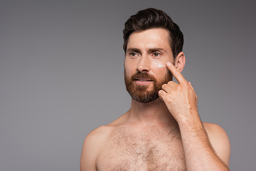 bearded and shirtless man applying cream on face isolated on grey