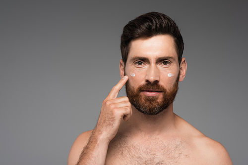 bearded man pointing with finger at cream on face isolated on grey