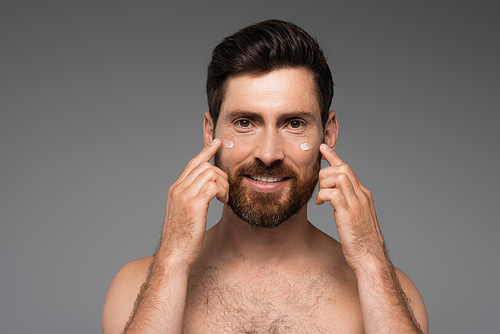 bearded man applying cream on face and smiling isolated on grey