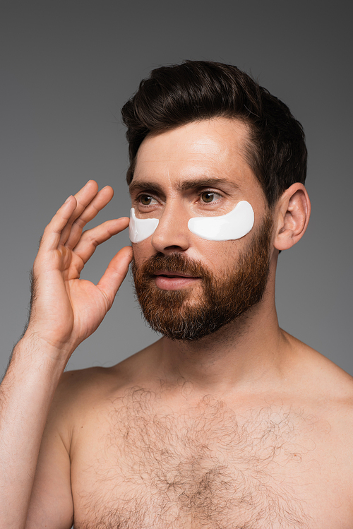 bearded man applying moisturizing eye patches isolated on grey