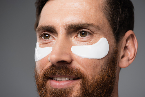 close up view of cheerful bearded man with moisturizing eye patches isolated on grey