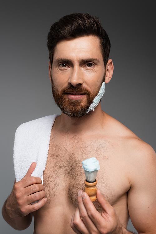 bearded man with shaving foam on face holding shaving brush isolated on grey