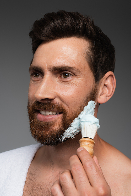 portrait of happy bearded man applying shaving foam with shaving brush isolated on grey
