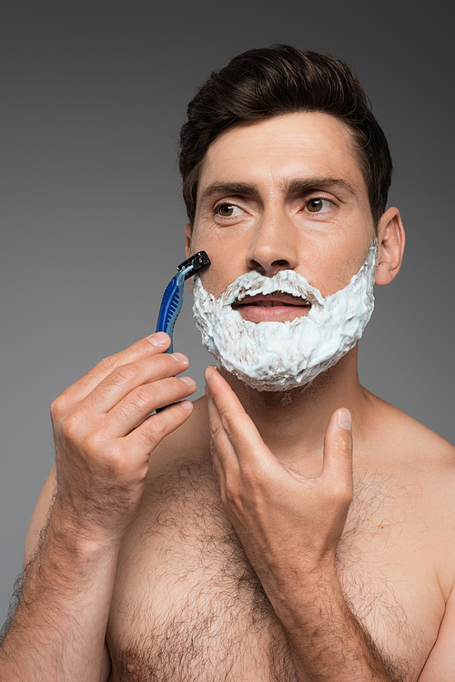 shirtless man with white shaving foam on face shaving with safety razor on grey