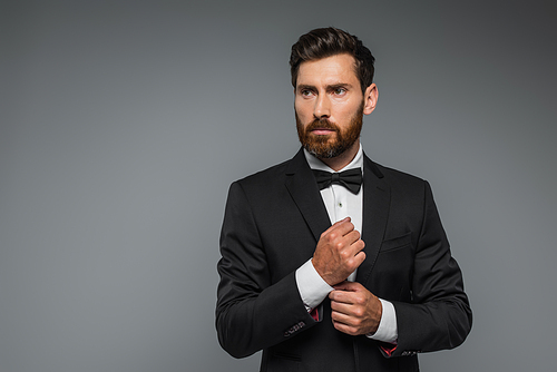 bearded man in black tuxedo with bow tie adjusting sleeve on white shirt isolated on grey