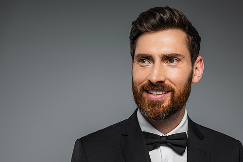 cheerful and bearded man in black tuxedo with bow tie isolated on grey