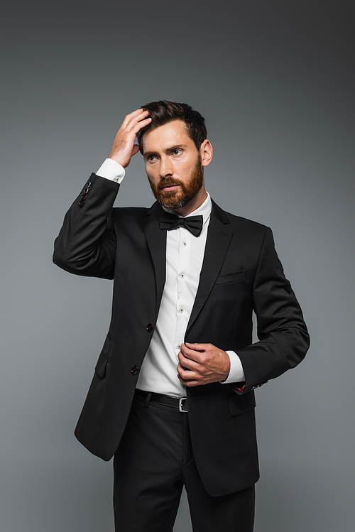 bearded man in black tuxedo with bow tie adjusting hair isolated on grey