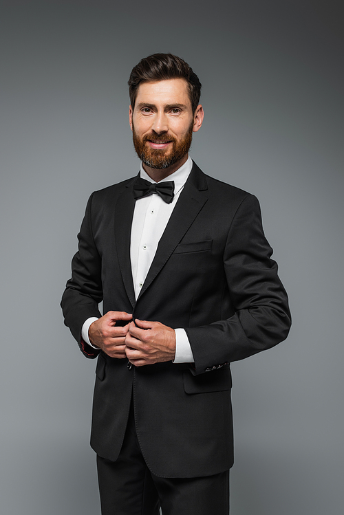 bearded man in black tuxedo with bow tie smiling isolated on grey