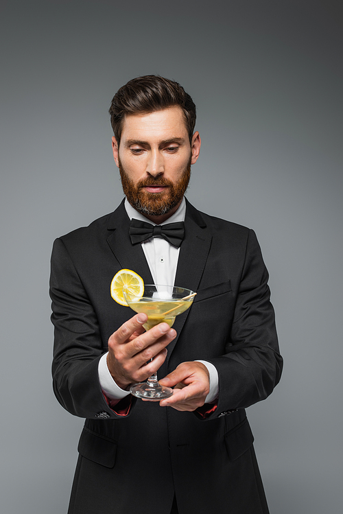 bearded man in elegant suit with bow tie holding glass with cocktail isolated on grey