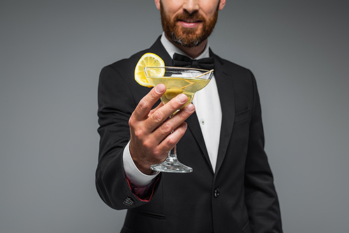 cropped view of cheerful and bearded man in suit with bow tie holding glass of cocktail isolated on grey