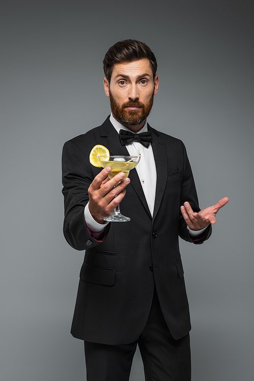 bearded man in elegant suit with holding glass with cocktail and gesturing isolated on grey