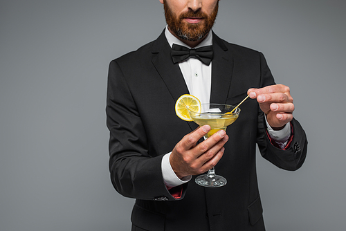partial view of bearded man in suit with bow tie holding toothpick with olive in glass with martini isolated on grey