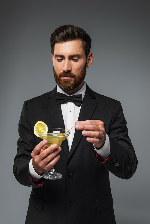 bearded man in suit with bow tie holding toothpick with olive in glass with martini isolated on grey