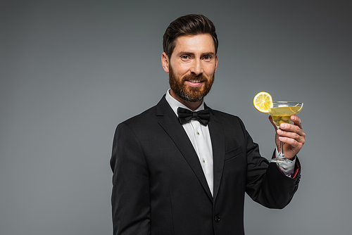 happy man in elegant suit with bow tie holding glass of cocktail isolated on grey