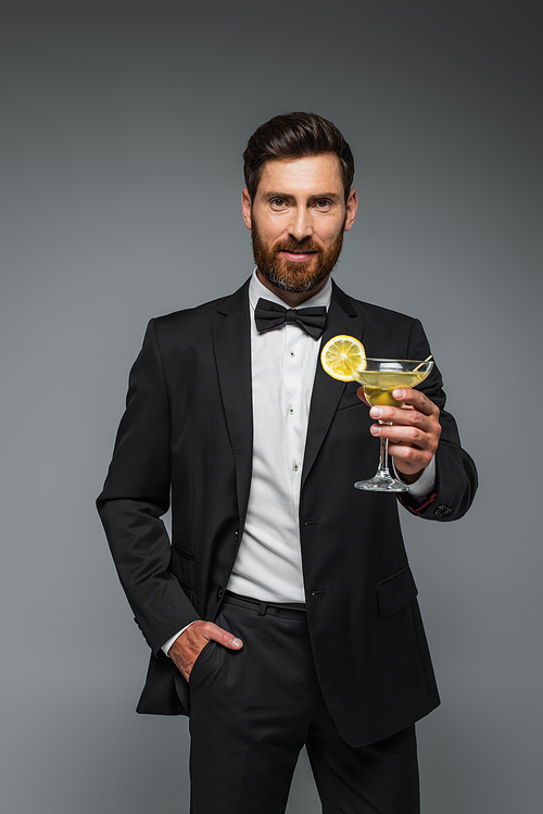 bearded man in elegant suit holding glass with cocktail and posing with hand in pocket isolated on grey