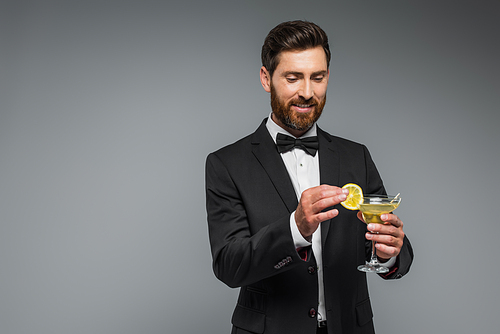 happy man in suit with bow tie holding sliced lemon and glass with cocktail isolated on grey