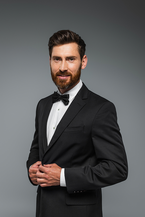 happy and successful man with beard standing in elegant suit with bow tie isolated on grey