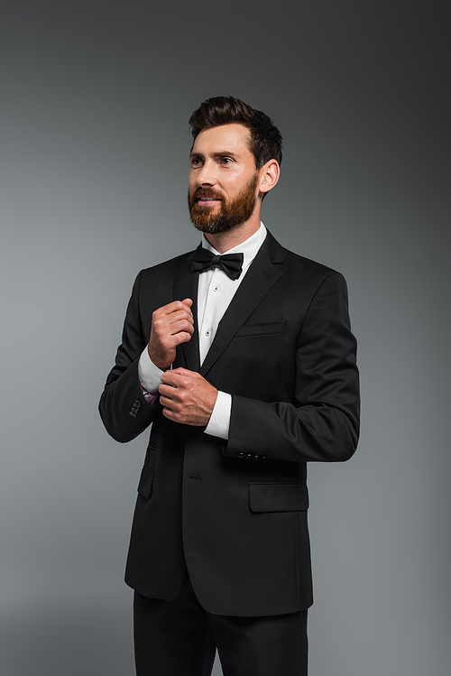 smiling man with beard standing in elegant suit with bow tie isolated on grey