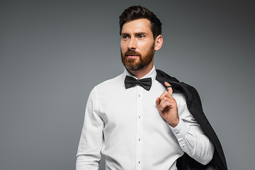 bearded man in white shirt with black bow tie holding blazer isolated on grey