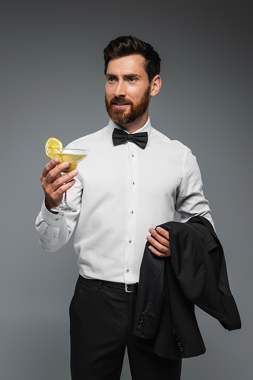 bearded man in tuxedo with bow tie holding glass with cocktail and blazer isolated on grey