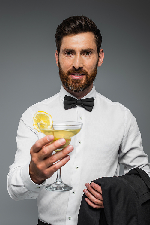 happy man in tuxedo with bow tie holding glass with cocktail and blazer isolated on grey