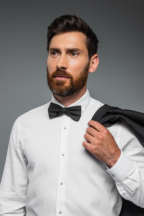 portrait of bearded man in suit with bow tie holding blazer isolated on grey