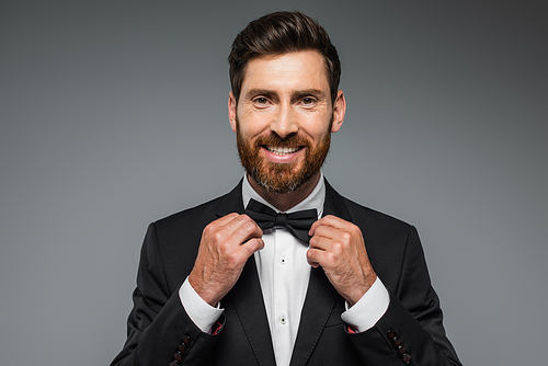 bearded man in suit smiling and adjusting bow tie isolated on grey