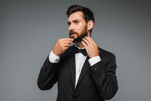man in tuxedo brushing beard with comb isolated on grey