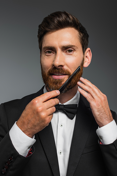 portrait of happy man in tuxedo brushing beard with comb isolated on grey