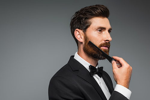 man in tuxedo brushing beard with comb and looking away isolated on grey