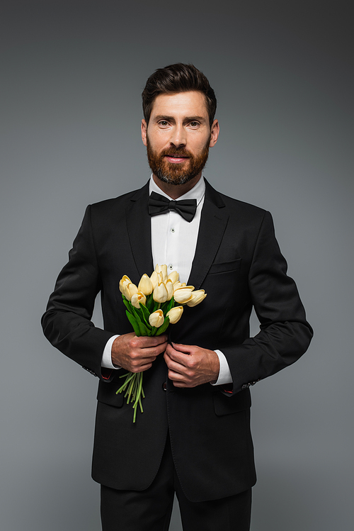 bearded man in elegant tux with bow tie holding bouquet of tulips isolated on grey
