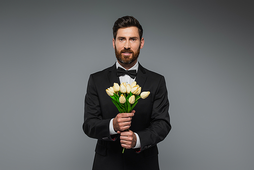bearded man in elegant suit with bow tie holding bouquet of tulips isolated on grey