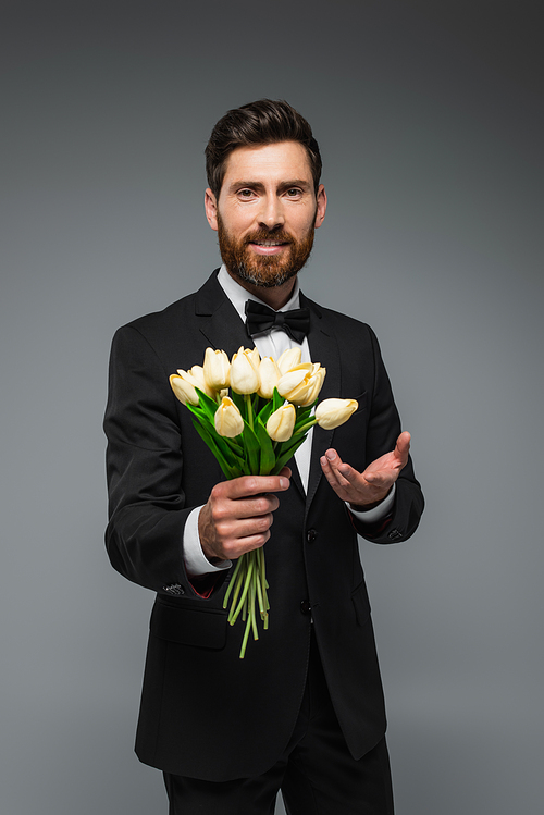 bearded man in elegant suit with bow tie holding fresh tulips and smiling isolated on grey