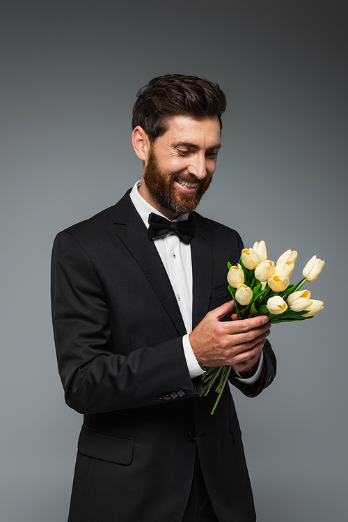 happy bearded man in elegant tuxedo with bow tie holding fresh tulips isolated on grey