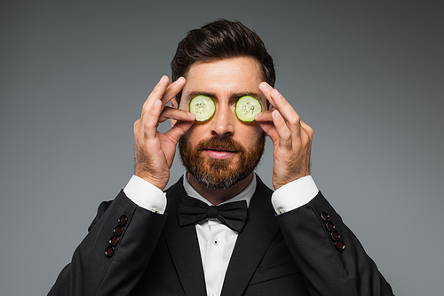 bearded man in tuxedo holding fresh sliced cucumber near eyes isolated on grey