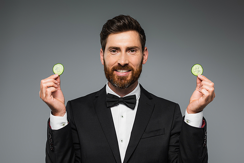 bearded man in tuxedo holding fresh sliced cucumber and smiling isolated on grey