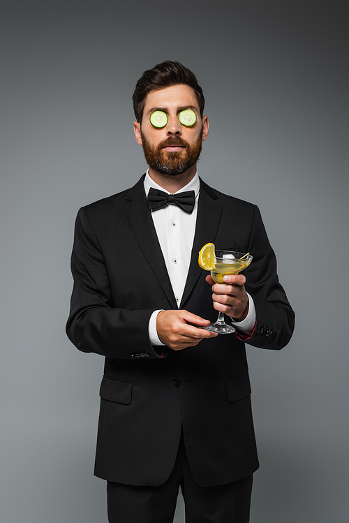 bearded man in tuxedo with fresh sliced cucumber on eyes holding glass of cocktail isolated on grey