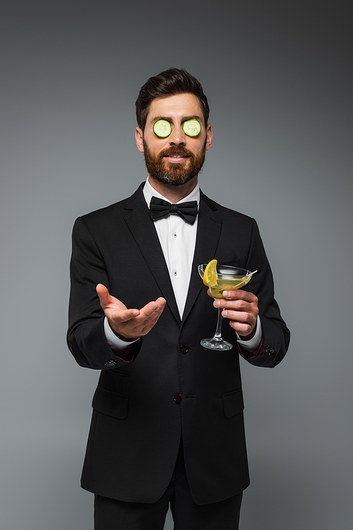 bearded man in tuxedo with fresh sliced cucumber on eyes holding cocktail isolated on grey