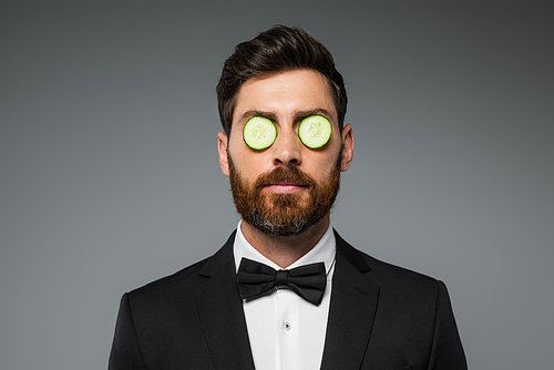 bearded man in tuxedo with fresh sliced cucumber on eyes isolated on grey