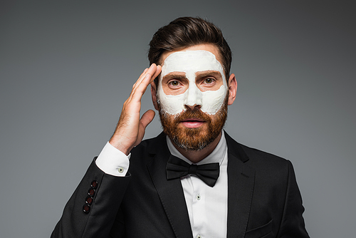 bearded man in suit with clay mask on face adjusting hair isolated on grey