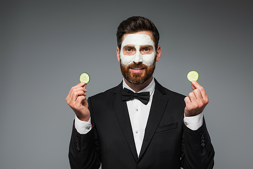 happy bearded man in suit with clay mask on face holding slices of cucumber isolated on grey