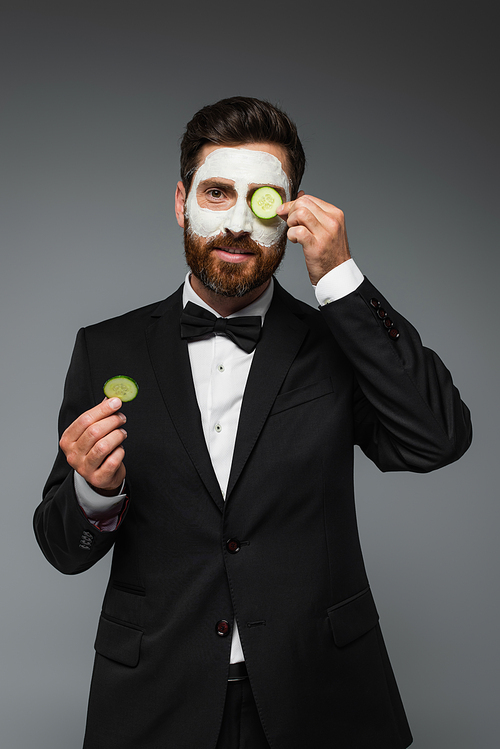 bearded man in suit with clay mask on face holding slices of cucumber isolated on grey