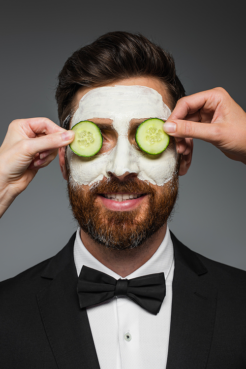 women applying sliced cucumbers on eyes of bearded man in suit with clay mask  isolated on grey