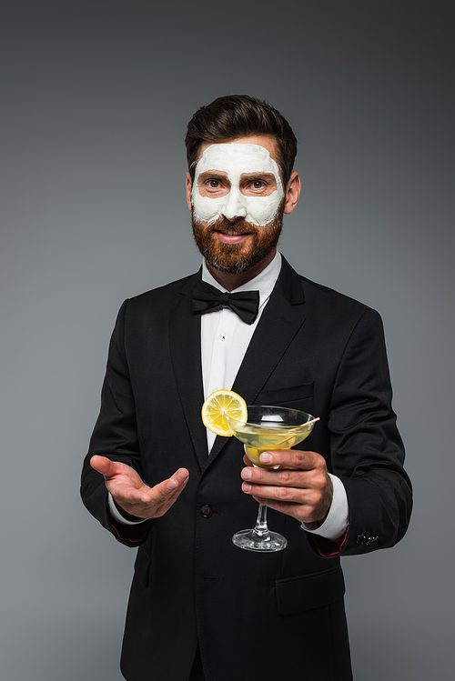 bearded man in tuxedo with clay mask on face holding cocktail and gesturing isolated on grey