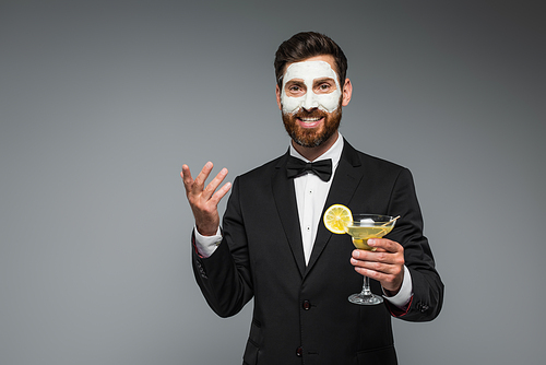joyful bearded man in suit with clay mask on face holding cocktail isolated on grey