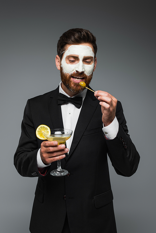 bearded man in suit with clay mask on face holding cocktail and toothpick with olive isolated on grey