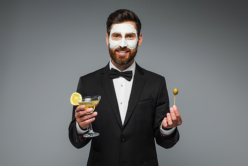 happy man in suit with clay mask on face holding cocktail and toothpick with olive isolated on grey