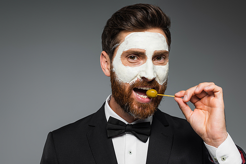bearded man in suit with clay mask on face holding toothpick and eating olive isolated on grey
