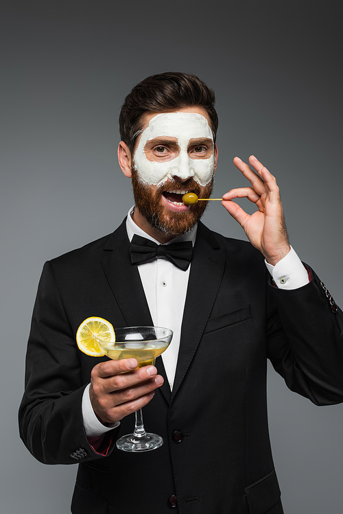 bearded man in suit with clay mask on face holding cocktail and eating olive isolated on grey